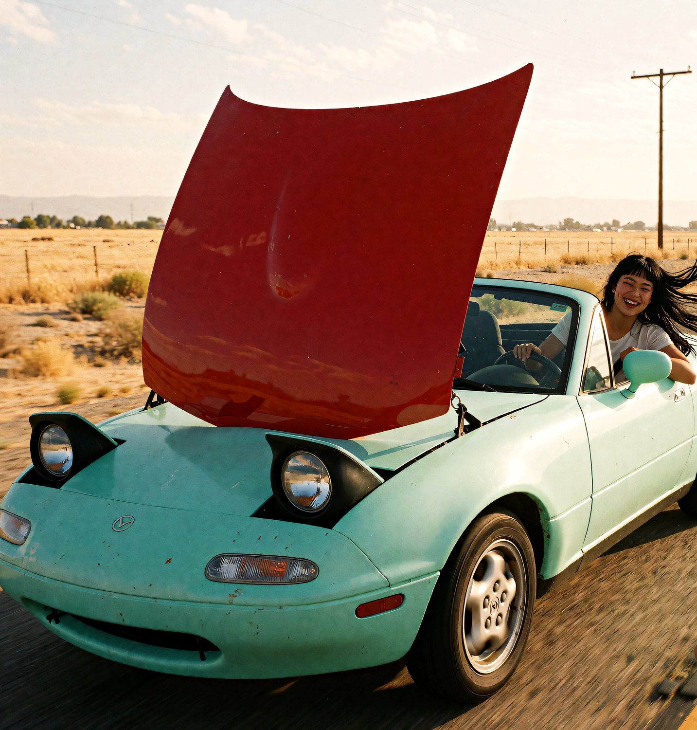 Juno laughs at the wheel of her all-mint Miata, a red car hood strapped upright on the front like a sail